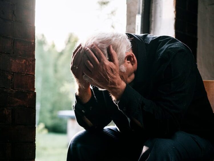 Senior man in black, grieving alone, sitting on a wooden chair by a doorway in a pensive pose.