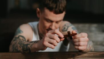 Tattooed man praying with a wooden crucifix inside a dimly lit church, symbolizing faith and reflection.