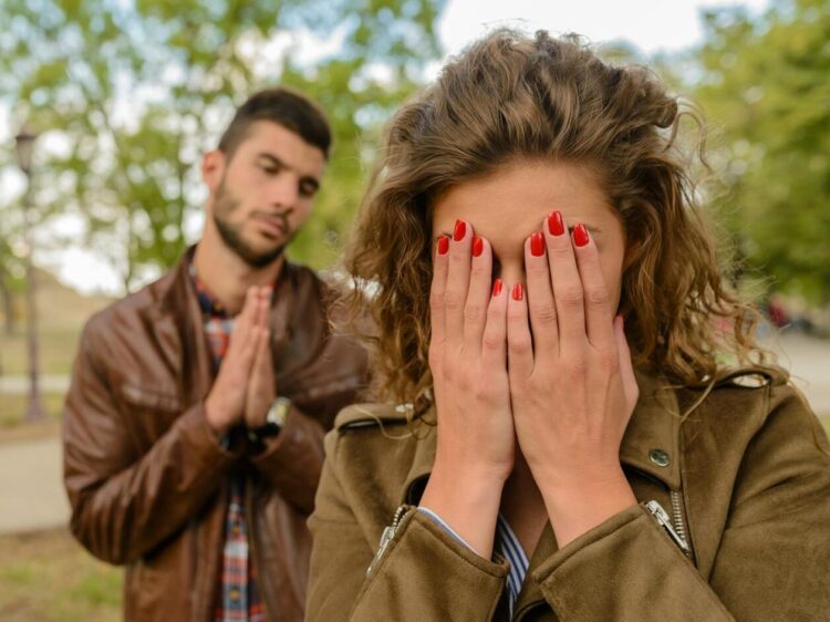 A young man apologizes while a woman covers her face, set in an outdoor park.