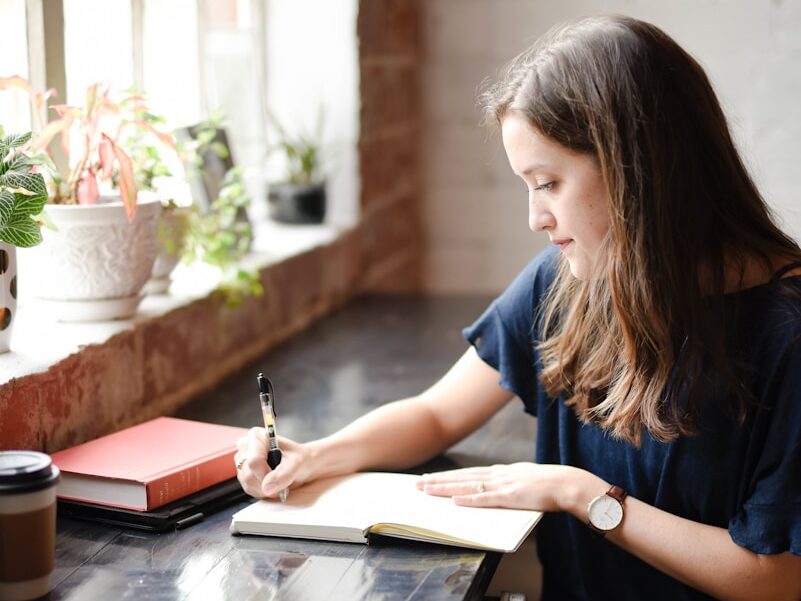 woman sitting in front of black table writing on white book near window
