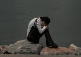 man in black and white jacket sitting on brown rock near body of water during daytime
