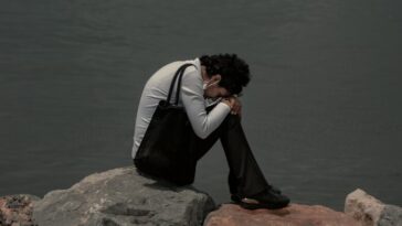man in black and white jacket sitting on brown rock near body of water during daytime