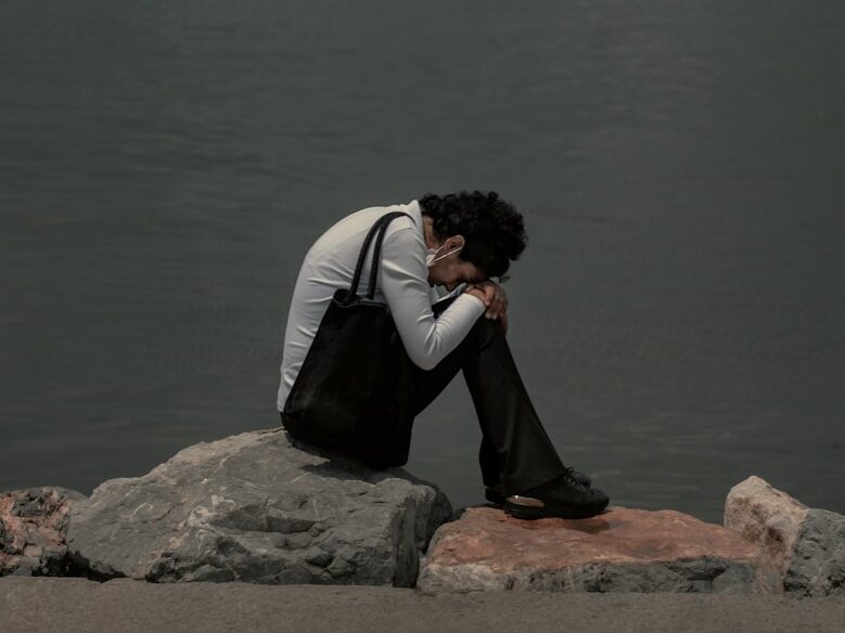 man in black and white jacket sitting on brown rock near body of water during daytime