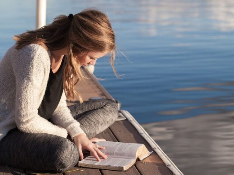 photo of woman reading book near body of water