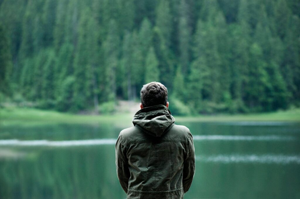A person in a green jacket stands by a serene forest lake, capturing calming nature.