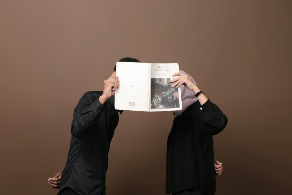 A man and woman in black holding a magazine, playfully hiding their faces.