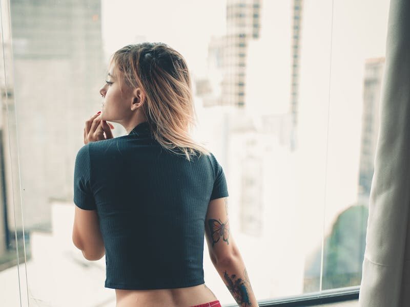 A thoughtful woman stands by a window, overlooking the cityscape in casual attire.