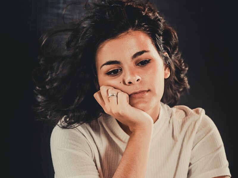 Portrait of a woman sitting pensively, wearing a casual white top. Moody, introspective atmosphere.