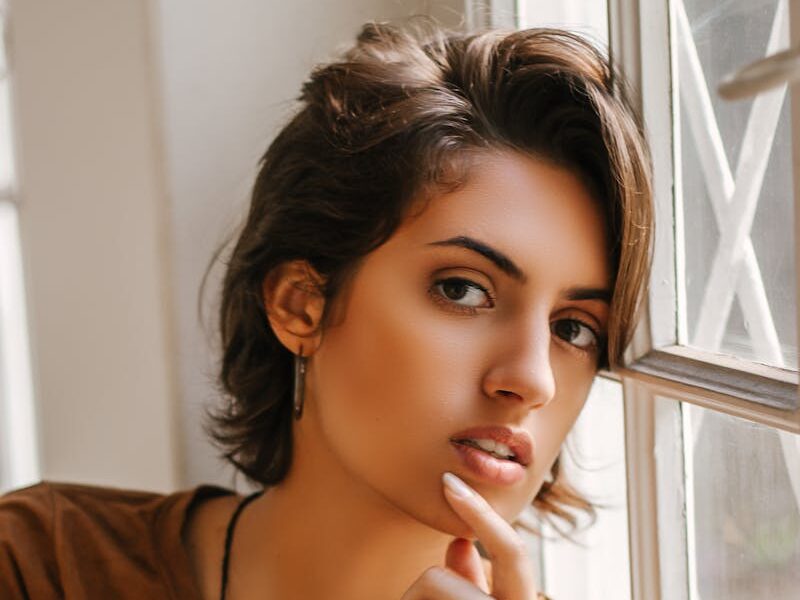 Graceful portrait of a thoughtful woman leaning against a window, capturing natural beauty and introspection.