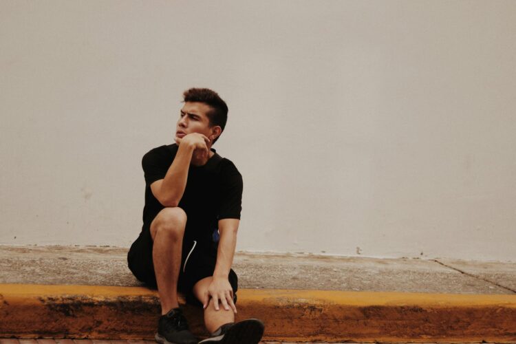 Man in casual black attire sitting thoughtfully on pavement against a white wall.