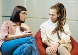 Two women engaged in a conversation in a modern office, sitting comfortably in relaxed chairs.