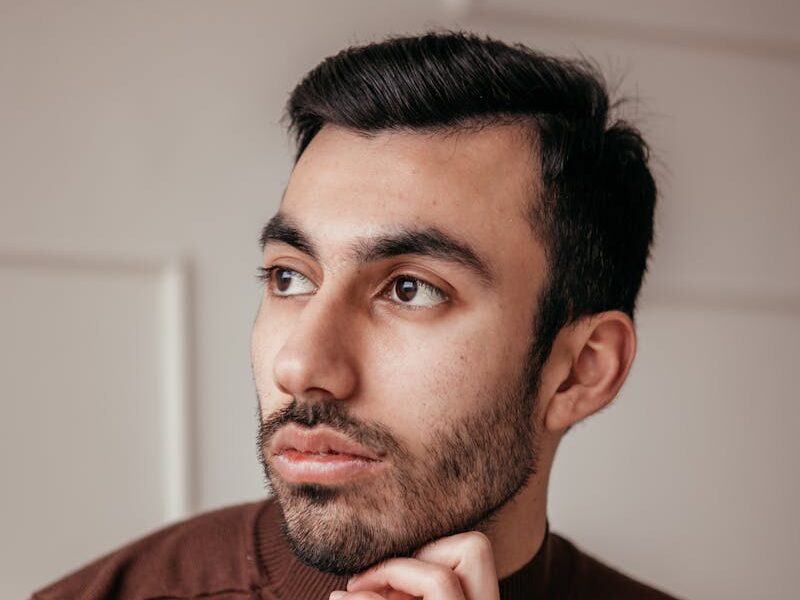 Portrait of a thoughtful young man wearing a brown sweater indoors, resting his chin on his hand.