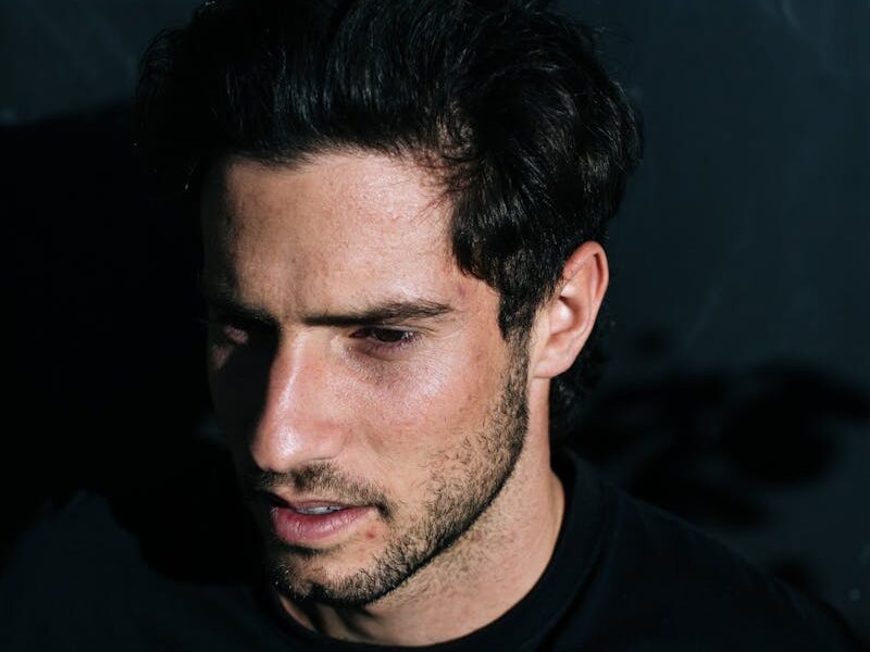 From above side view of pensive male with beard and dark hair looking away while standing on black background in bright sunshine