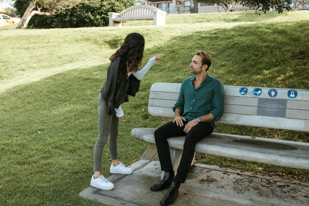 A couple in a heated argument while sitting and standing in a sunny park setting.