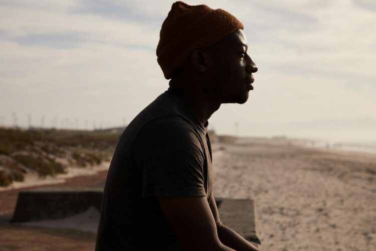 Side view of African American male in hat sitting on cement border on sandy shore near sea against cloudy sky