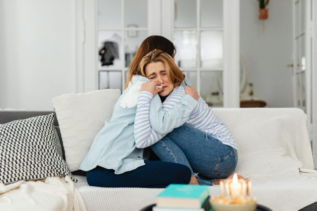 Two women embracing on a sofa, providing comfort and support in a warm, well-lit living room.