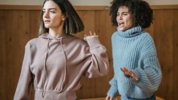 Mad African American female in warm sweater screaming at irritated female while having argument in light room with wooden walls