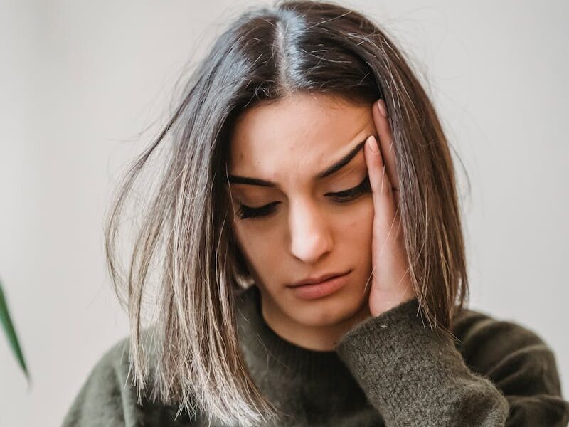 Unhappy female in casual wear leaning on hand and surfing internet on cellphone while sitting in light room near wall at home