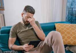 A man sits on a sofa holding a tablet, visibly stressed and covering his face with one hand.
