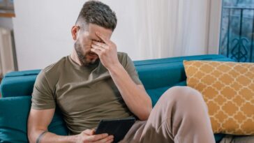 A man sits on a sofa holding a tablet, visibly stressed and covering his face with one hand.