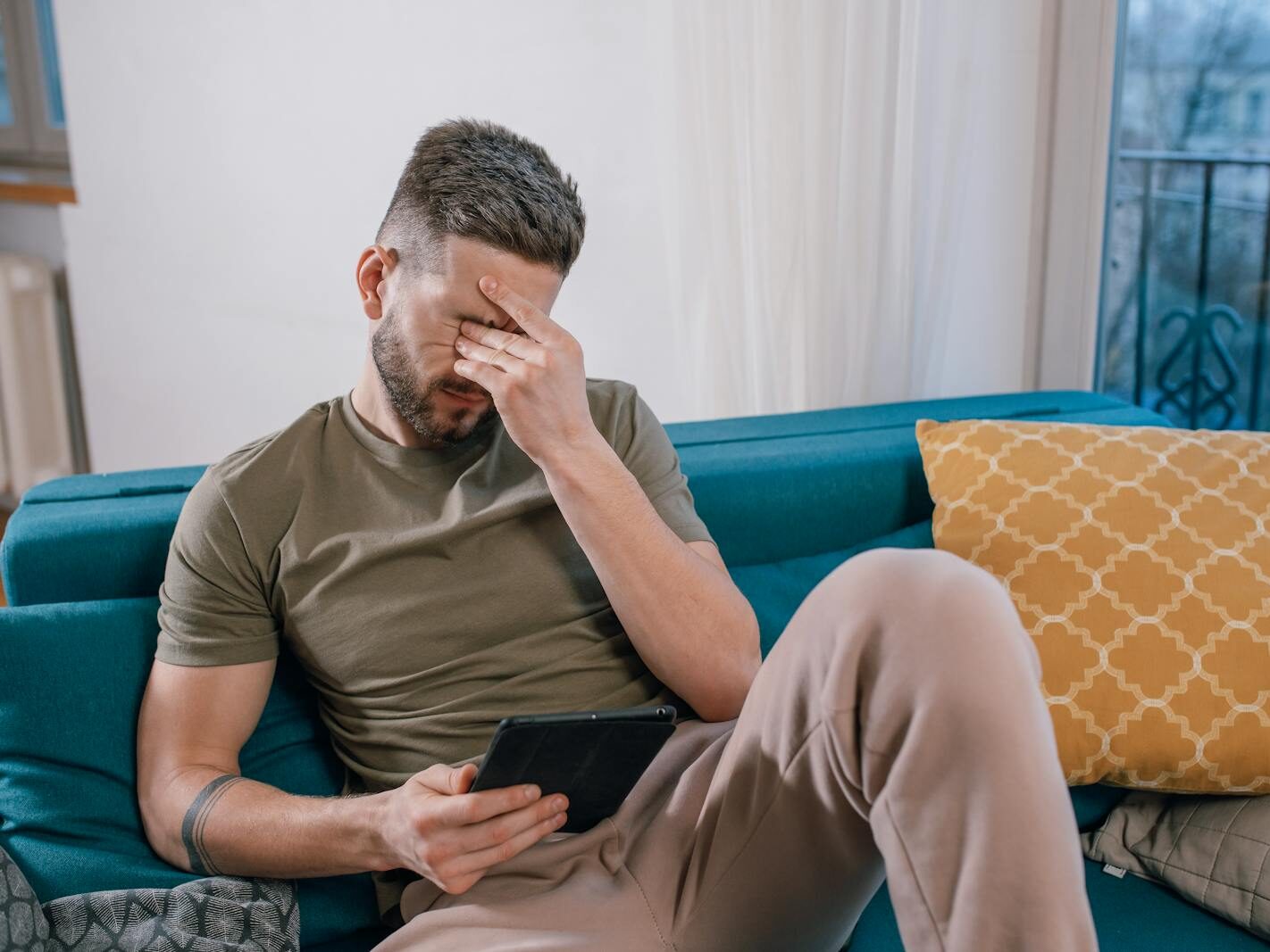 A man sits on a sofa holding a tablet, visibly stressed and covering his face with one hand.