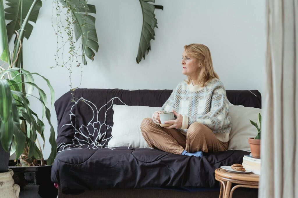 Full body of peaceful middle aged woman with blond hair in casual clothes drinking coffee and looking away thoughtfully while resting on sofa with crossed legs