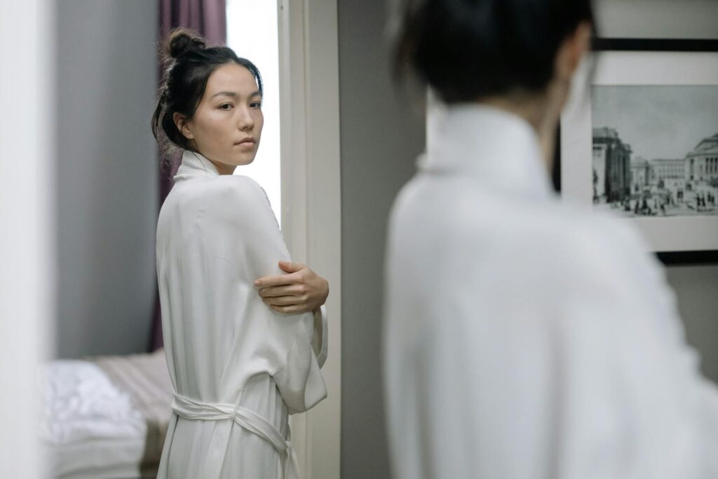 Woman in a white bathrobe gazing at her reflection in the mirror indoors.