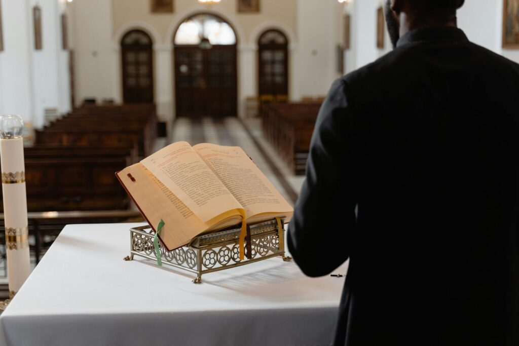 A priest reads a Bible on a stand inside a serene church interior with empty pews.
