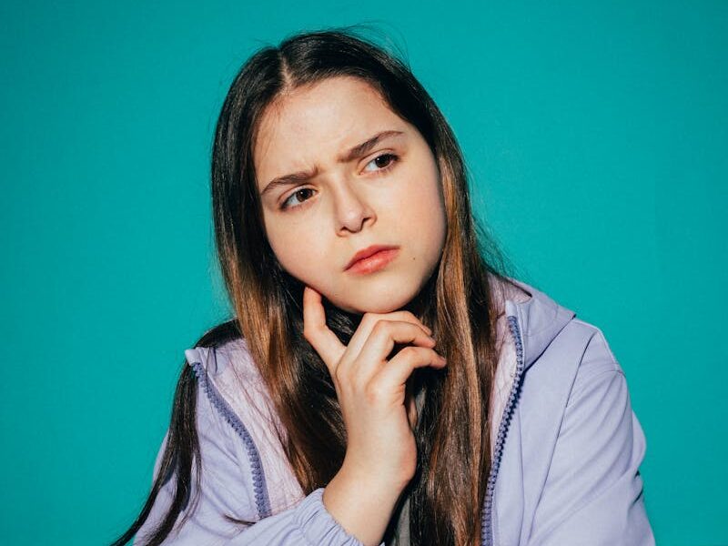 A young girl pondering deeply against a vibrant teal background.