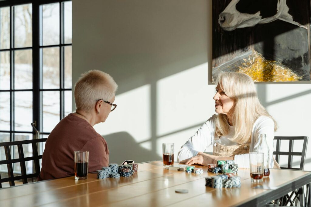 Two senior women sitting at a table playing poker with drinks, enjoying leisure time indoors.