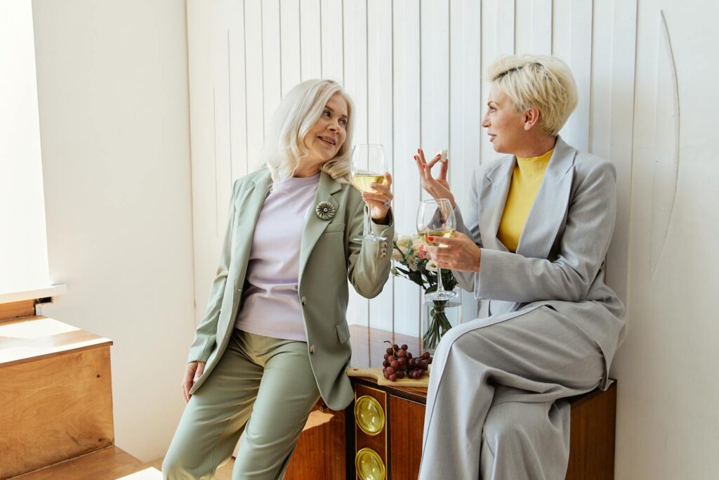 Two stylish mature women in suits enjoy wine indoors, engaged in cheerful conversation.