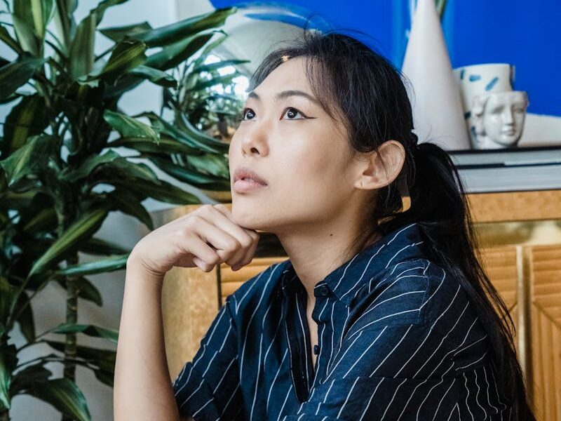 Asian woman sitting thoughtfully indoors beside indoor plants.