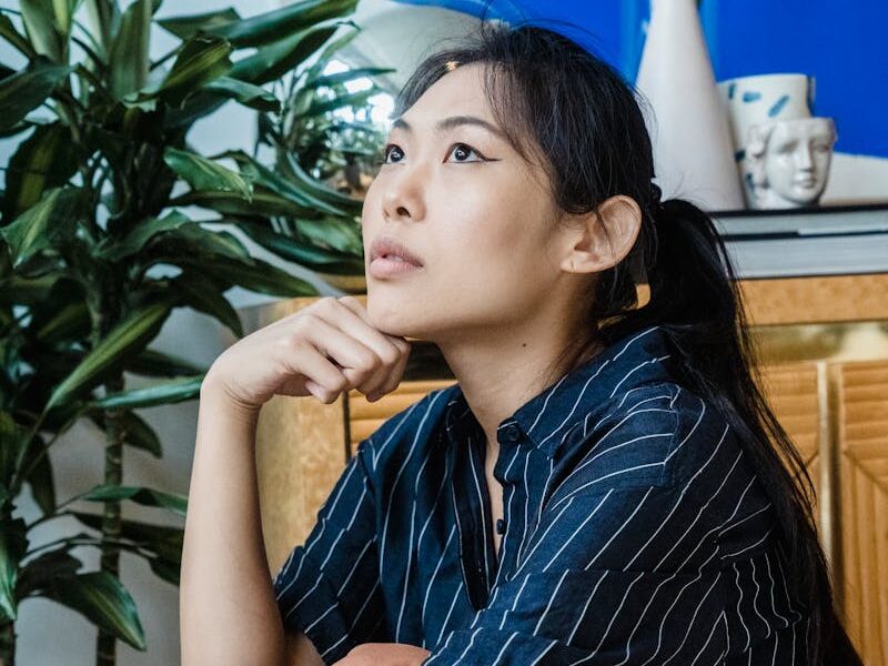 Asian woman sitting thoughtfully indoors beside indoor plants.