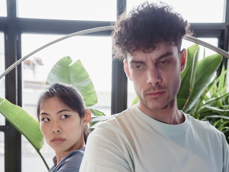 Couple standing back to back with crossed arms and tense expressions, surrounded by indoor plants.