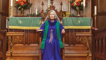 Elderly woman in clergy attire sitting on a wooden chair in a church, surrounded by altar decorations.