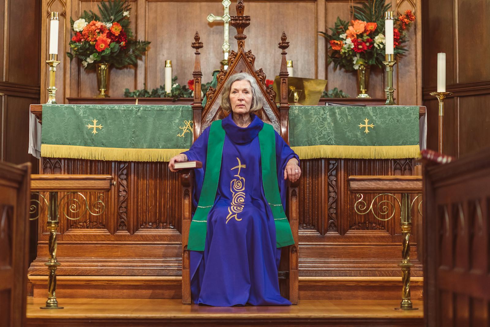 Elderly woman in clergy attire sitting on a wooden chair in a church, surrounded by altar decorations.