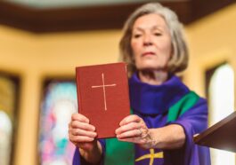 An elderly female priest holding a Bible inside a church setting.