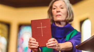 An elderly female priest holding a Bible inside a church setting.