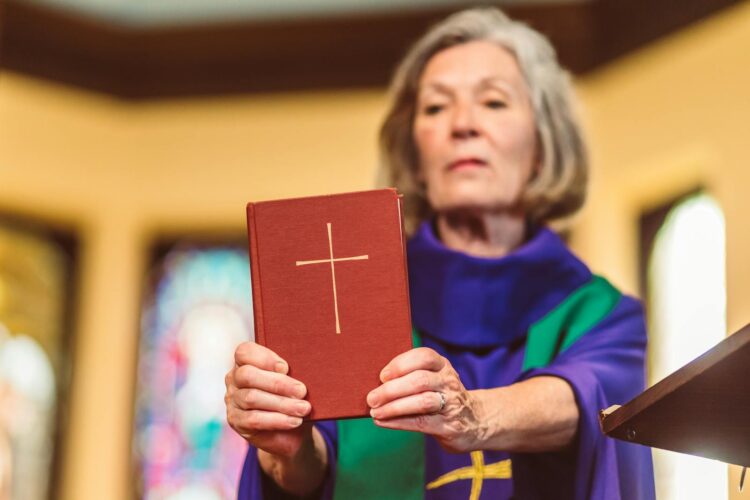 An elderly female priest holding a Bible inside a church setting.