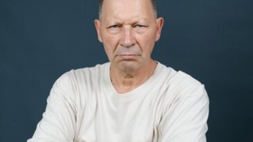 Bald middle-aged man wearing a white t-shirt posing with crossed arms against a gray background.
