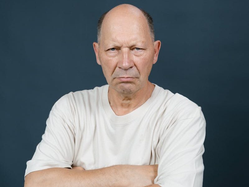 Bald middle-aged man wearing a white t-shirt posing with crossed arms against a gray background.