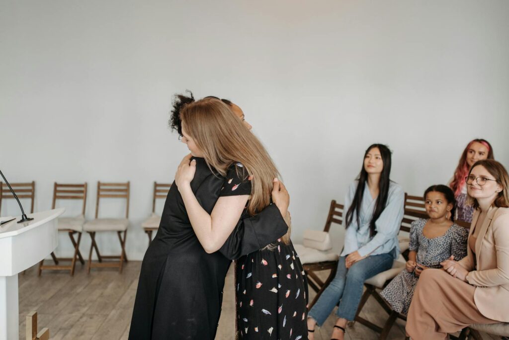 Diverse group of women sharing an embrace indoors while others observe. A sense of community and support is depicted.