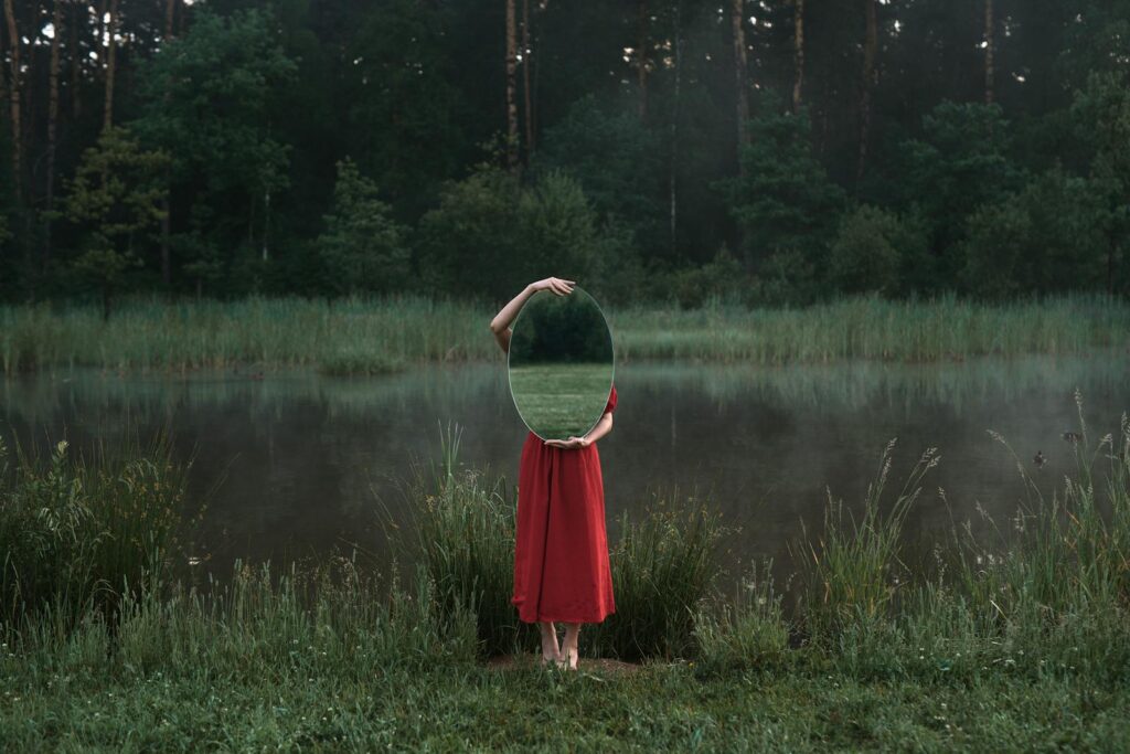 Artistic photo of a woman in nature holding a mirror beside a misty pond.