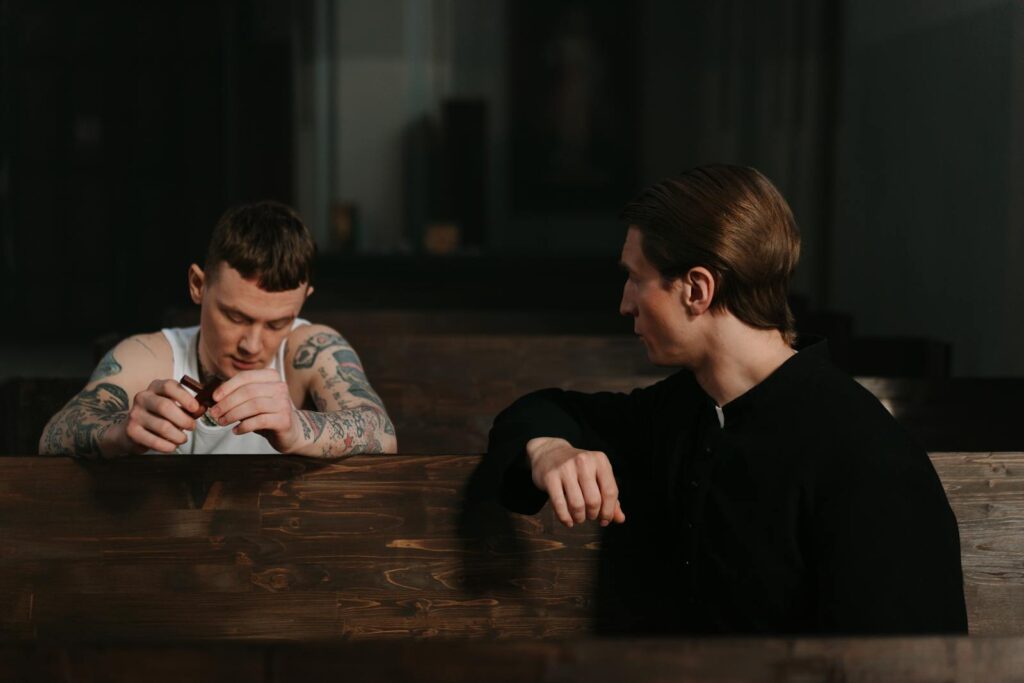 A tattooed man in conversation with a priest inside a church, sharing a moment of confession.