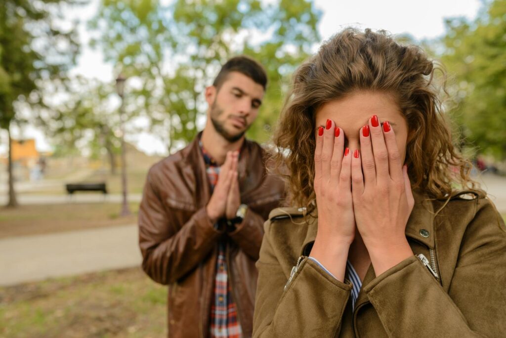 A young man apologizes while a woman covers her face, set in an outdoor park.