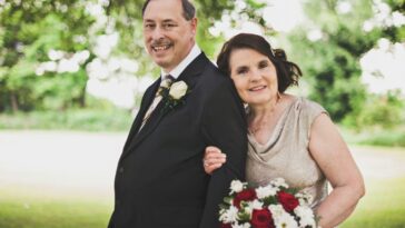 man in black suit standing beside woman in gray sleeveless dress holding bouquet of flowers