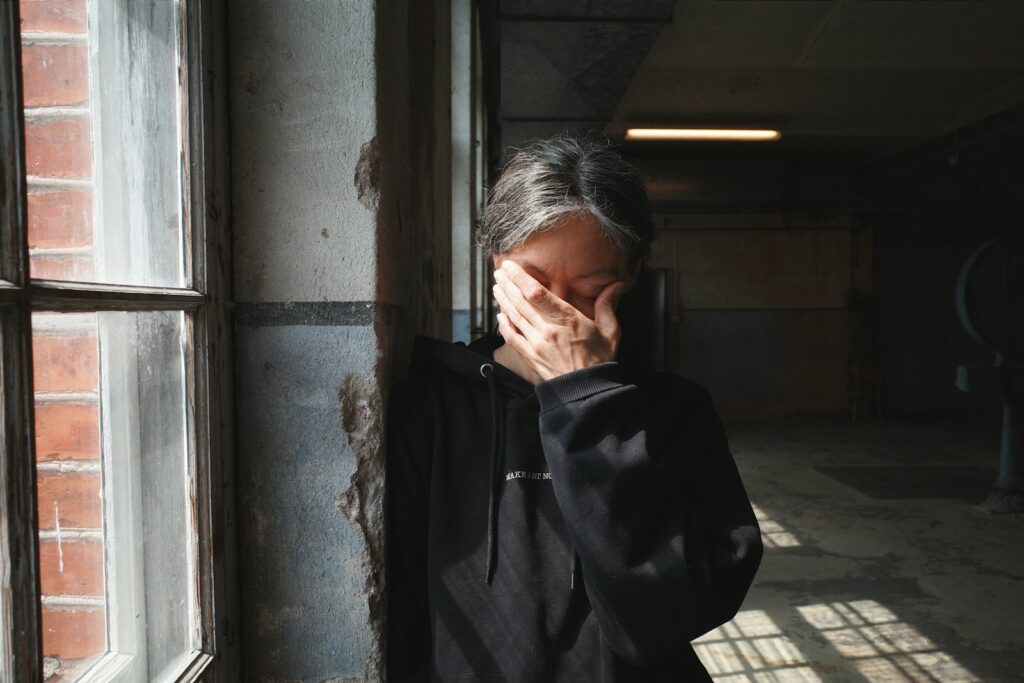 Person covers face with hand near window.