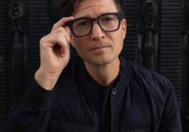 a man wearing glasses sitting in front of a black wall