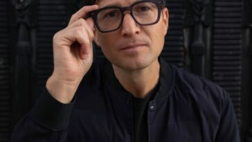 a man wearing glasses sitting in front of a black wall