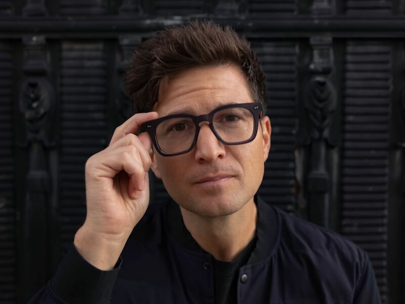 a man wearing glasses sitting in front of a black wall